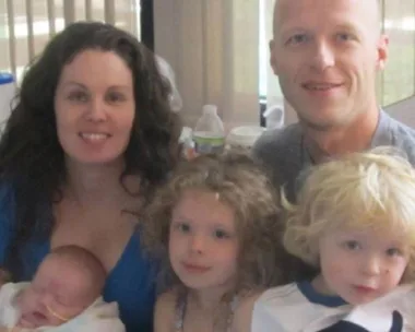 A family of five, including a mother, father, and three young children, pose together indoors, smiling.