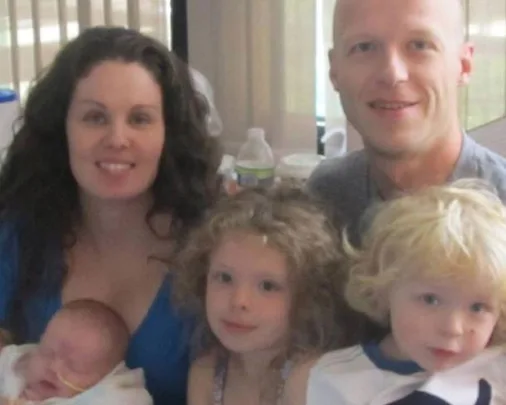 A family of five, including a mother, father, and three young children, pose together indoors, smiling.