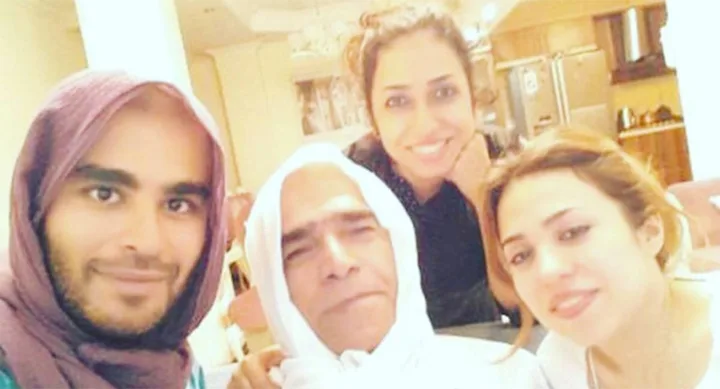 Group of four people smiling indoors, with some wearing headscarves, in a cozy setting with kitchen in the background.