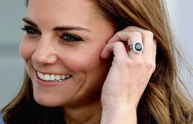 A woman smiling, showing a large sapphire and diamond ring on her left hand.