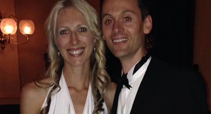 A couple dressed formally, smiling at a social event with warm lighting in the background.