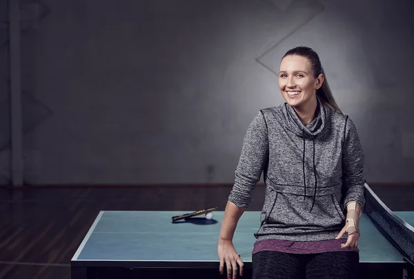 A woman smiles while sitting on a table tennis table, wearing a gray hoodie in a dimly lit room.
