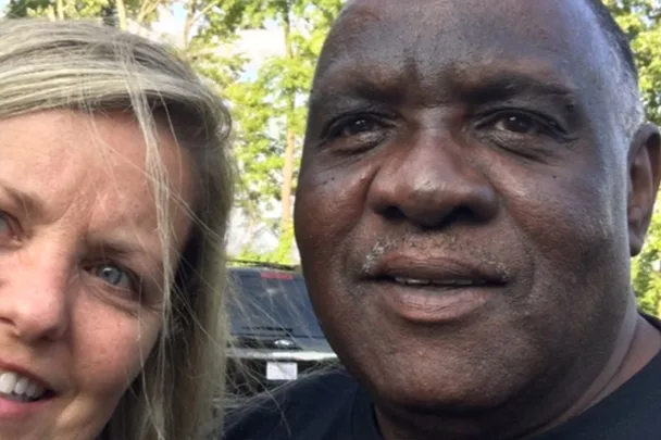 A close-up of a smiling man and woman, both looking directly at the camera, with trees and a car in the background.