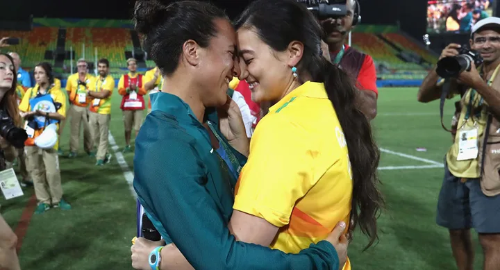 Two athletes hugging and smiling with their foreheads touching, surrounded by photographers and cheering spectators at the Rio Olympics.