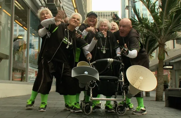 Group of elderly people wearing matching outfits and sneakers, posing energetically on a city street with a stroller and megaphone.