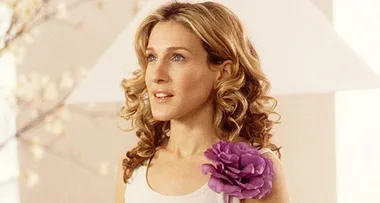 A woman with curly blonde hair, wearing a white top with a large purple flower on her shoulder, looking off to the side.