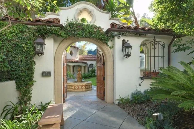Spanish-style villa entrance with an arched wooden gate, surrounded by greenery, leading to a courtyard with a fountain.