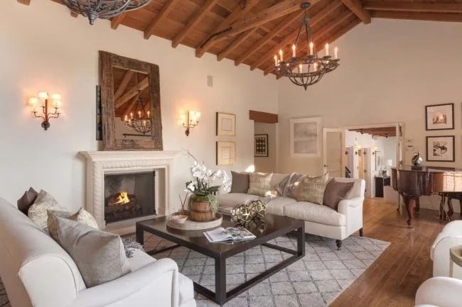 Luxurious living room with white sofas, fireplace, wooden ceiling, chandelier, and decor on a geometric-patterned rug.