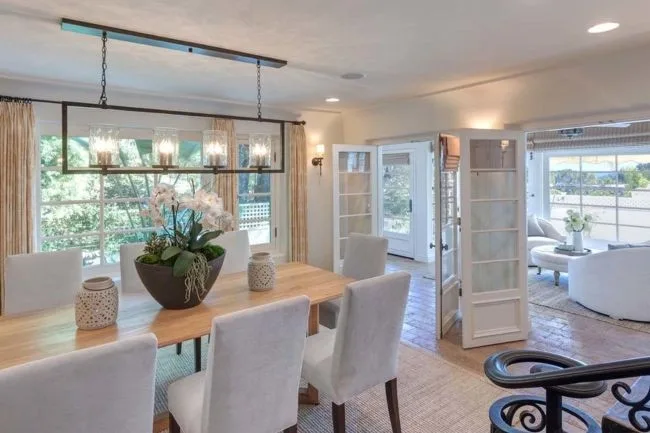 Dining area with wooden table, white chairs, chandelier, large plant centerpiece, and open doors to a sunlit room.
