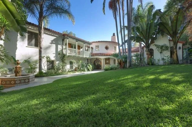 Spanish-style villa with lush green lawn, palm trees, and tiled roof, featuring a fountain and white balconies.