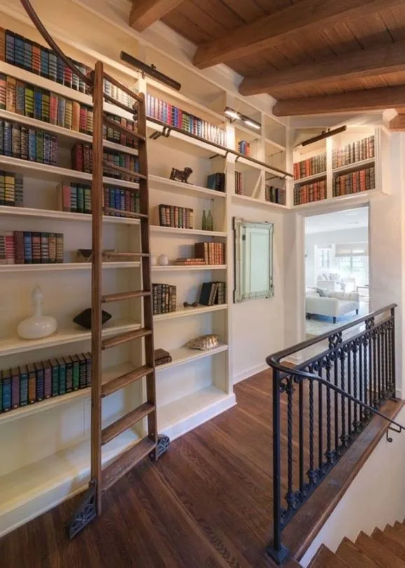 Library with wooden ladder, built-in bookshelves full of colorful books, and a view into a living room.