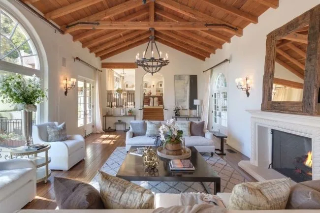Spacious living room with high wooden ceiling, chandelier, fireplace, white sofas, large windows, and a central coffee table.