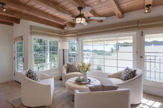 Cozy sunlit living room with white armchairs, round ottoman, and large windows overlooking a scenic view.