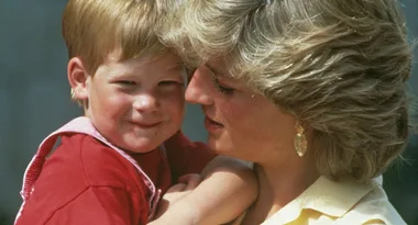 A woman with short blonde hair lovingly holds a smiling child with red hair wearing a red shirt and overalls.