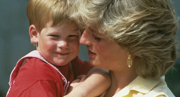 A woman with short blonde hair lovingly holds a smiling child with red hair wearing a red shirt and overalls.