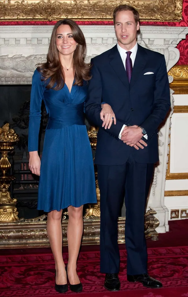 Two people in formal attire stand together, the woman in a blue dress and the man in a dark suit, in an ornate room.