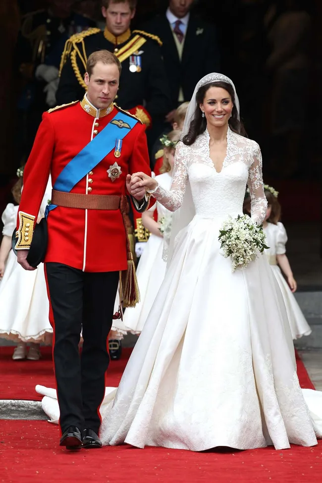 Royal couple on wedding day; bride in lace gown, groom in red uniform, holding hands on red carpet, smiling, with floral bouquet.