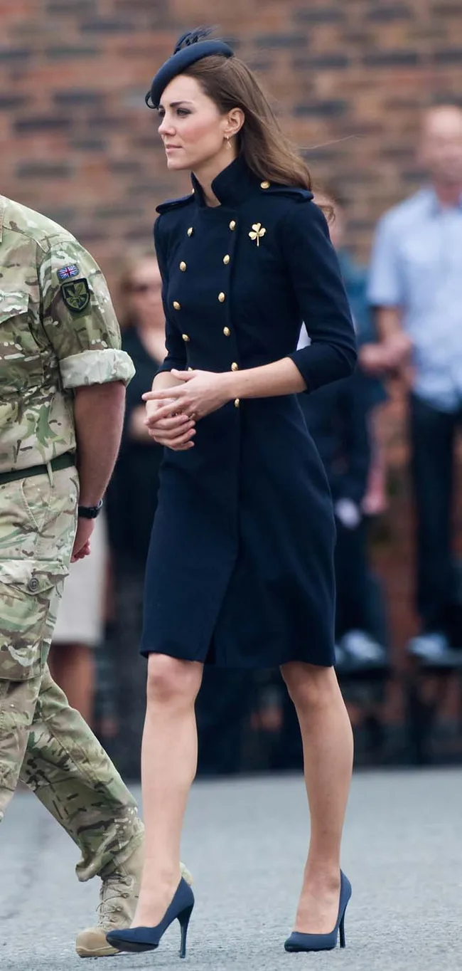 A woman in a navy military-style coat, hat, and heels walks beside a person in camouflage attire.