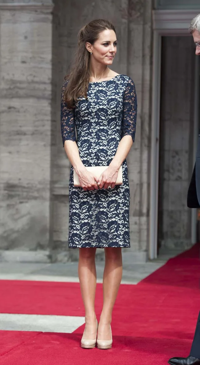 A person in a navy lace dress on a red carpet, holding a beige clutch, standing beside an ornate building entrance.