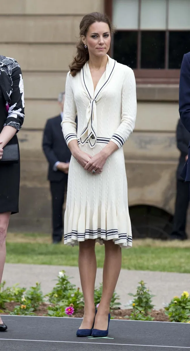 A woman in a cream dress with black trim stands outdoors, surrounded by others, on a sunny day.