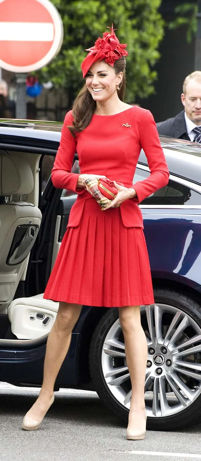 A woman in a red dress and hat stands beside a car, holding a patterned clutch, smiling.