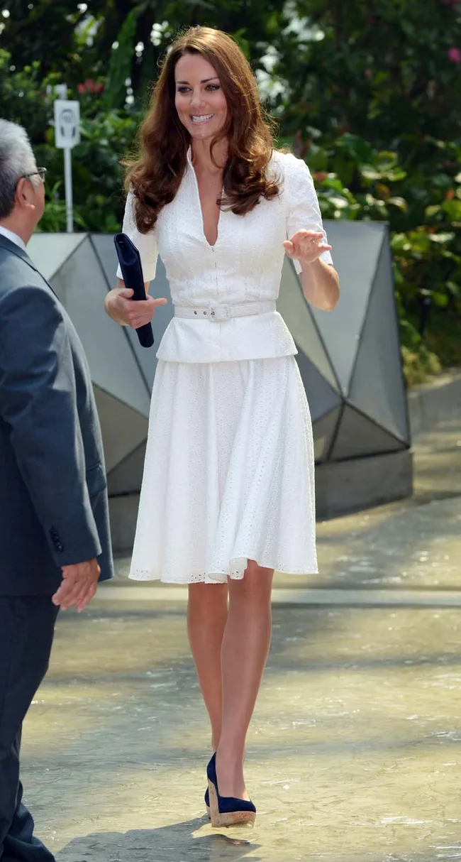 A woman in a white dress and navy wedges smiling while walking outside on a sunny day.