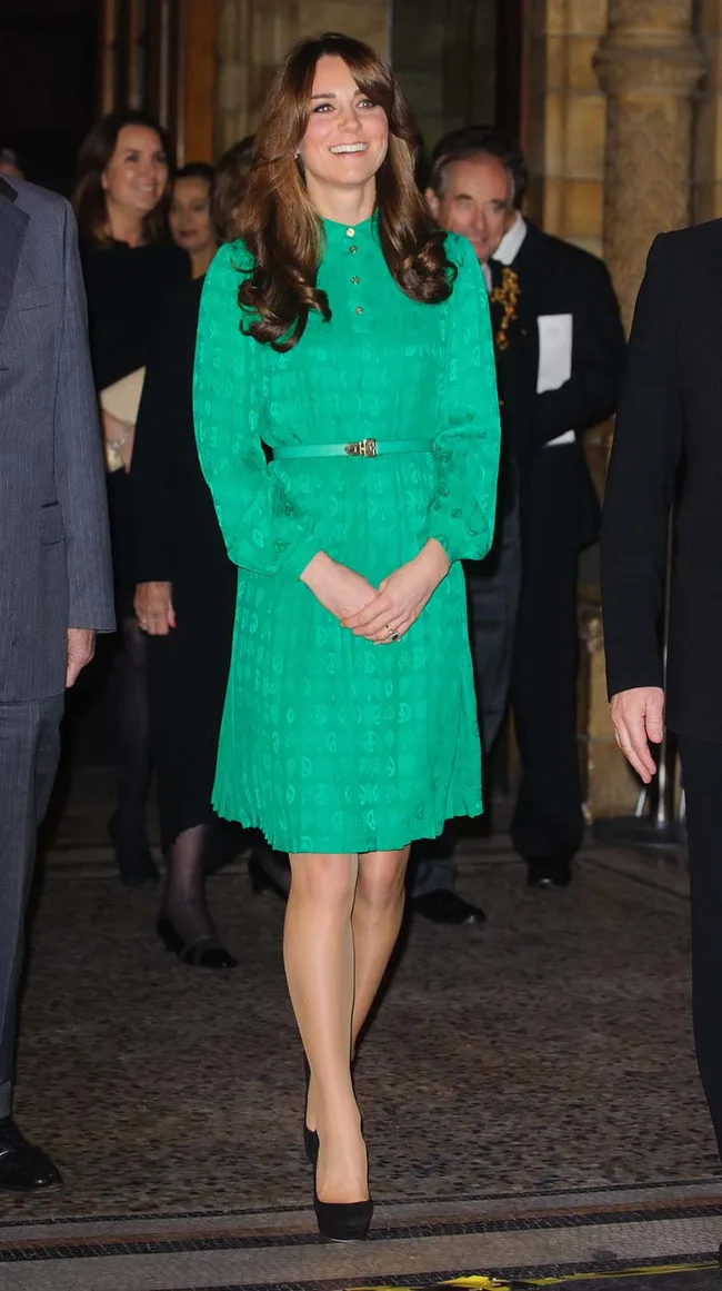 A woman in a green dress with long brown hair smiles while standing indoors, surrounded by people.