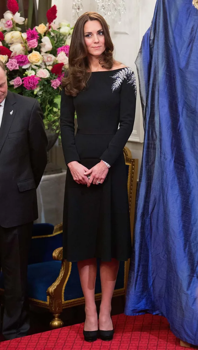 A woman in a black dress with silver embellishment on her shoulder, standing near a chair with floral decor in the background.
