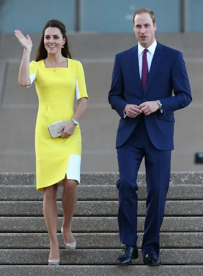A woman in a yellow dress waves while walking down steps with a man in a blue suit.