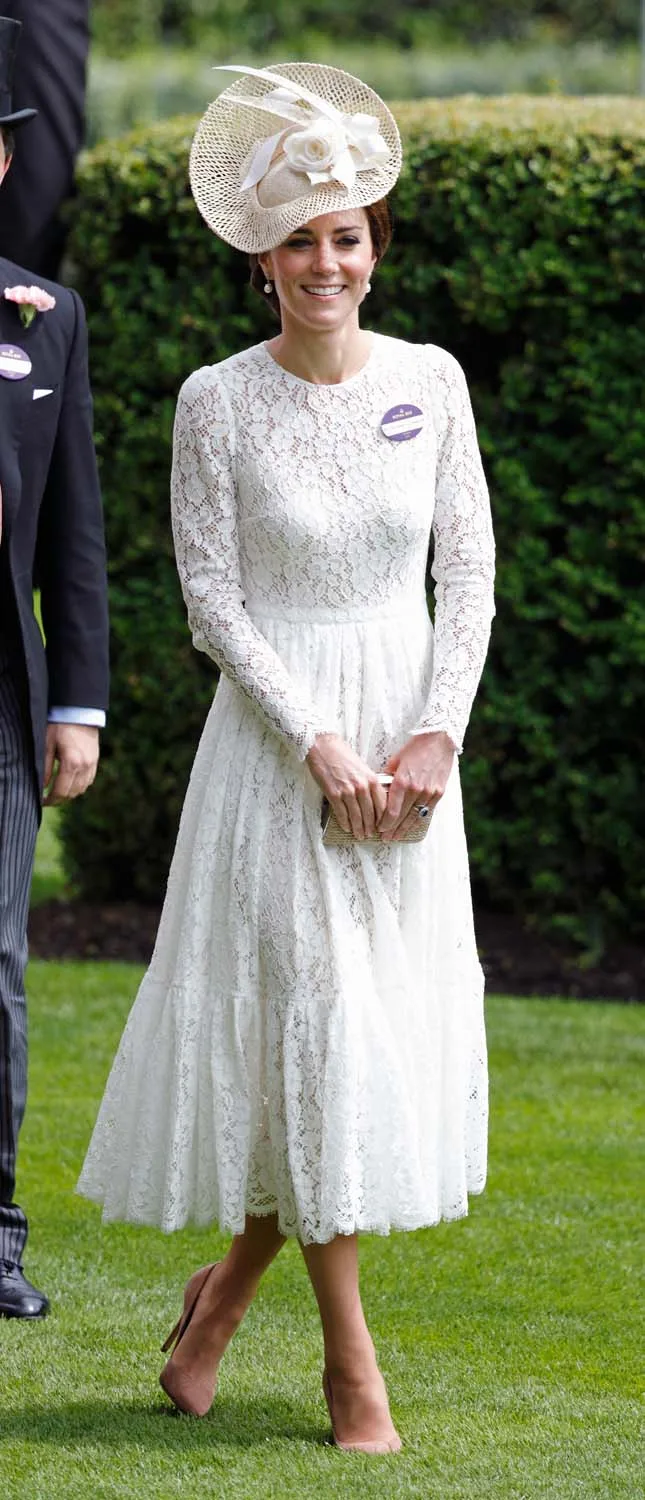 A woman in a white lace dress and large hat stands on grass, smiling.