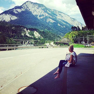 A person sitting barefoot on a roadside, holding a child, with a backdrop of mountains under a partly cloudy sky.