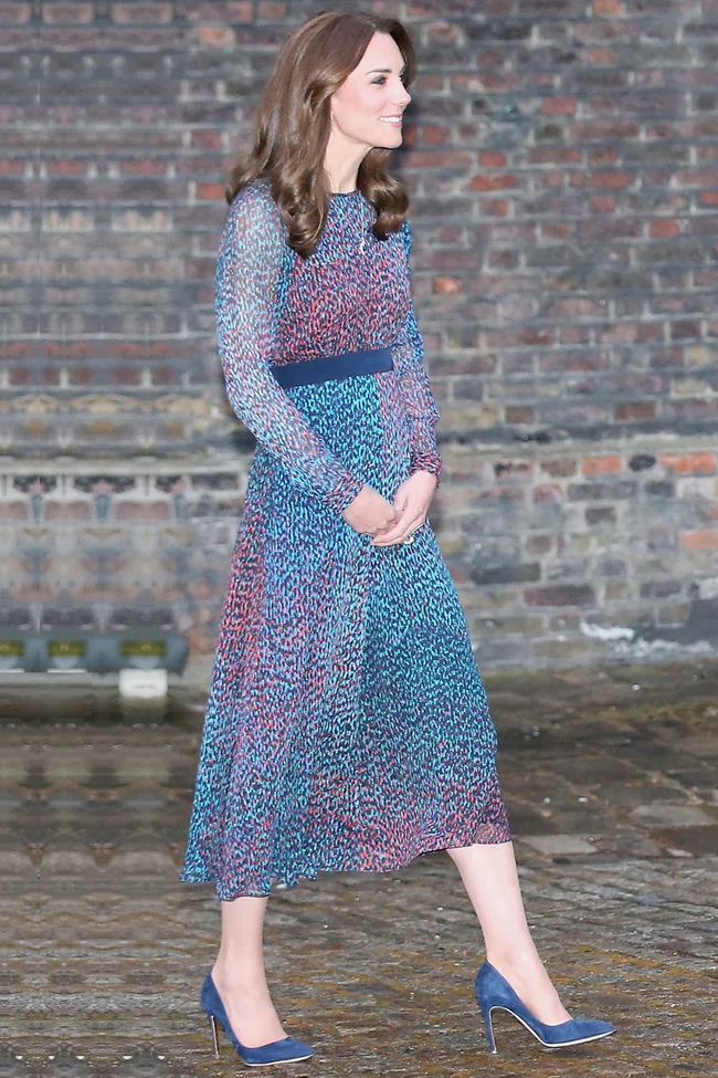 A person in a multicolored dress and blue heels stands on a cobblestone path with a brick wall background.