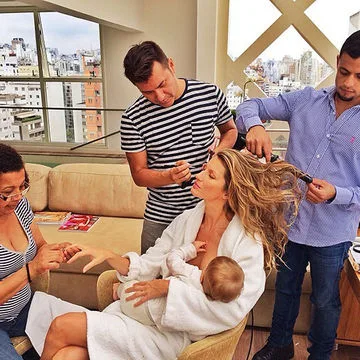 Woman in robe getting hair and nails done while breastfeeding, assisted by a team, in a city-view room.
