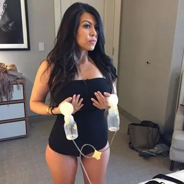 Woman using a breast pump in a hotel room, wearing a black top, surrounded by bags and a framed photo on the wall.