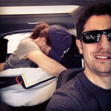 Man taking a selfie in a car, wearing sunglasses; woman in the back resting on the car seat.