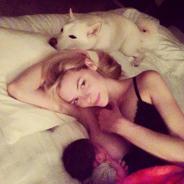 A woman lying on a bed with a baby and a white dog beside her.