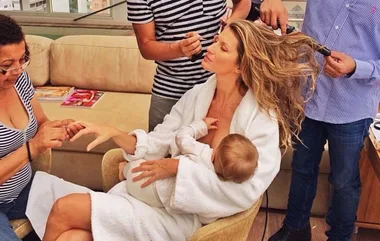 A woman in a robe breastfeeds a baby while getting her hair styled and nails done by two people.