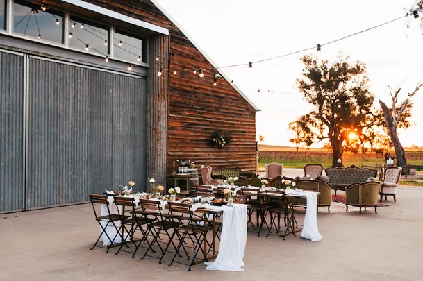 Outdoor rustic wedding reception setup with wooden tables and vintage furniture under string lights at sunset.