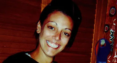 A woman smiling in front of wooden blinds, with colorful patterns on the right side.