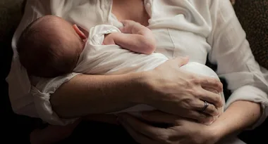 Mother holding a newborn baby in her arms, wearing a white shirt, with gentle lighting.
