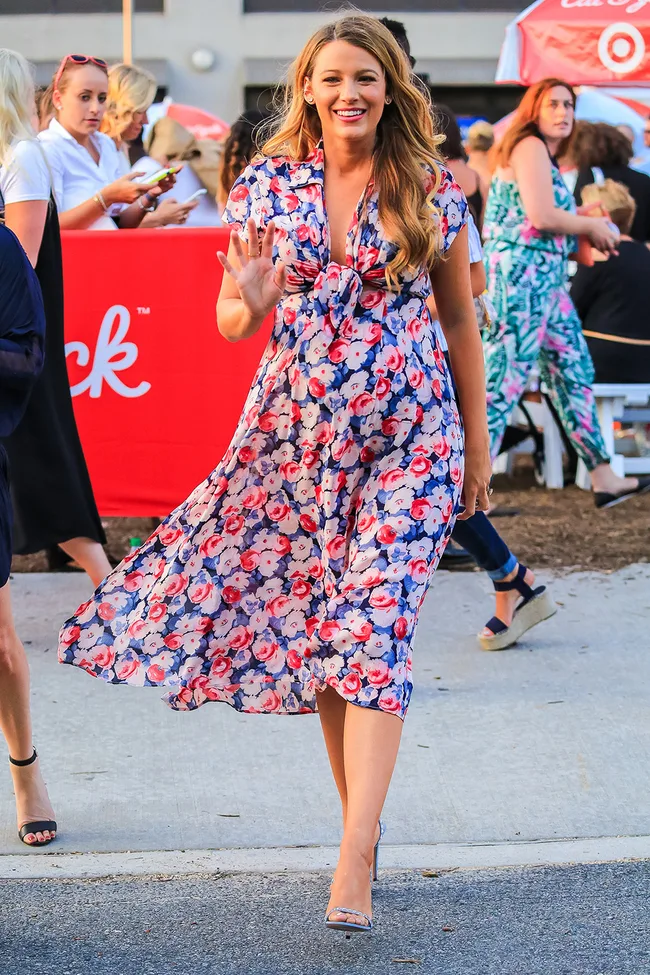Woman in a floral dress waves while walking outside, surrounded by people in a busy outdoor setting.