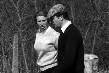 Black-and-white photo of a man in a flat cap walking with a woman in a sweater, with trees in the background.