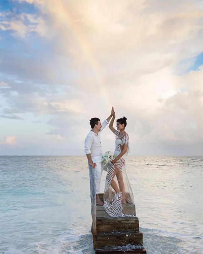 Couple in white clothing stands on a wooden pier above the ocean, under a colorful sky with a faint rainbow.