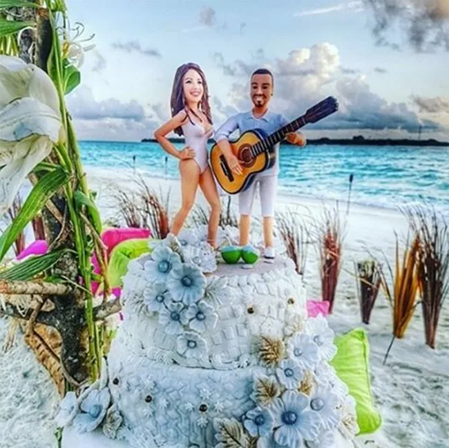 Cake with beach-themed figurines of a woman in a swimsuit and a man playing guitar on a beach with ocean background.