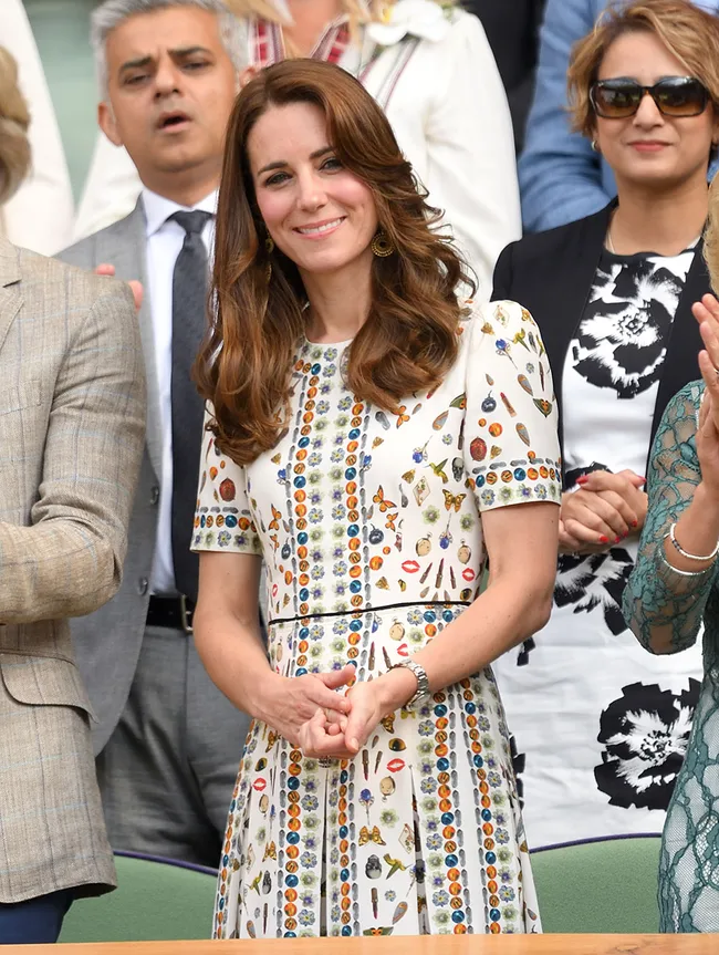A woman in a white patterned dress smiles, standing among others in a crowd, with a man and woman behind her.