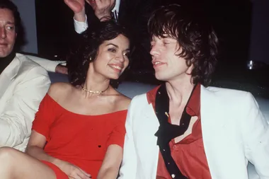 A woman in a red dress smiles at a man in a white suit and red shirt, both seated in a dimly lit room.