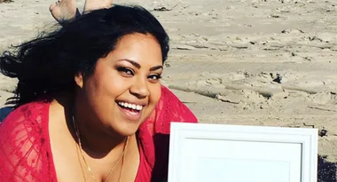 Smiling person in a red dress on a sandy beach holding a picture frame.
