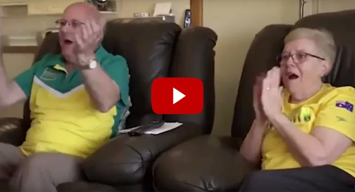 Elderly couple in matching sports jerseys cheer excitedly from their recliners.