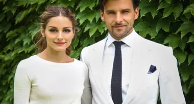 A woman in a white dress and a man in a white suit pose in front of green leaves.