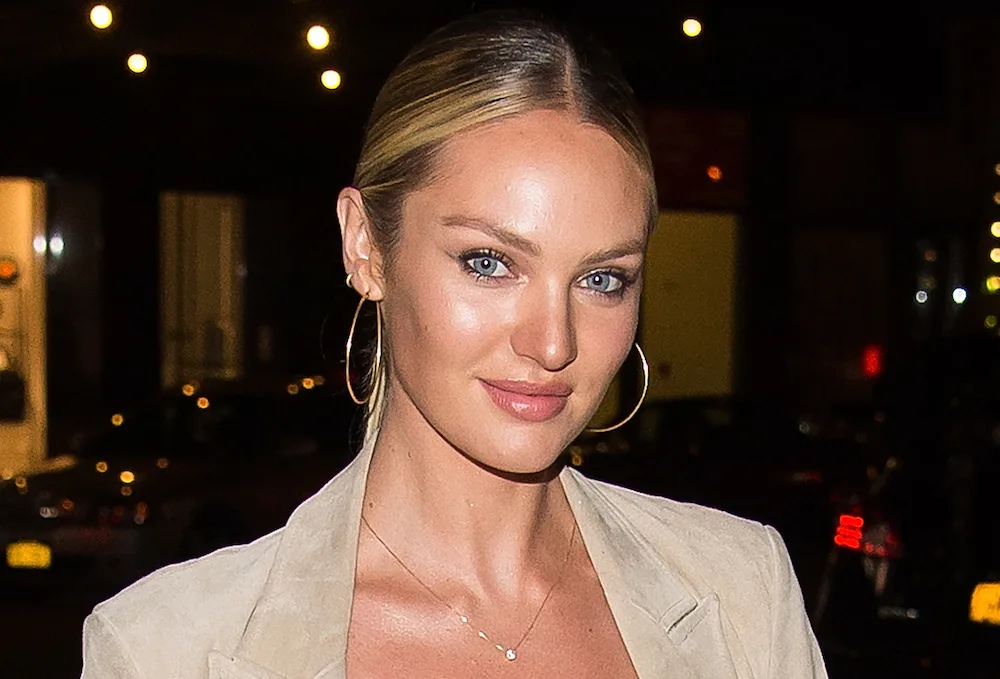 A person with blonde hair and hoop earrings smiles at night, wearing a light-colored outfit.